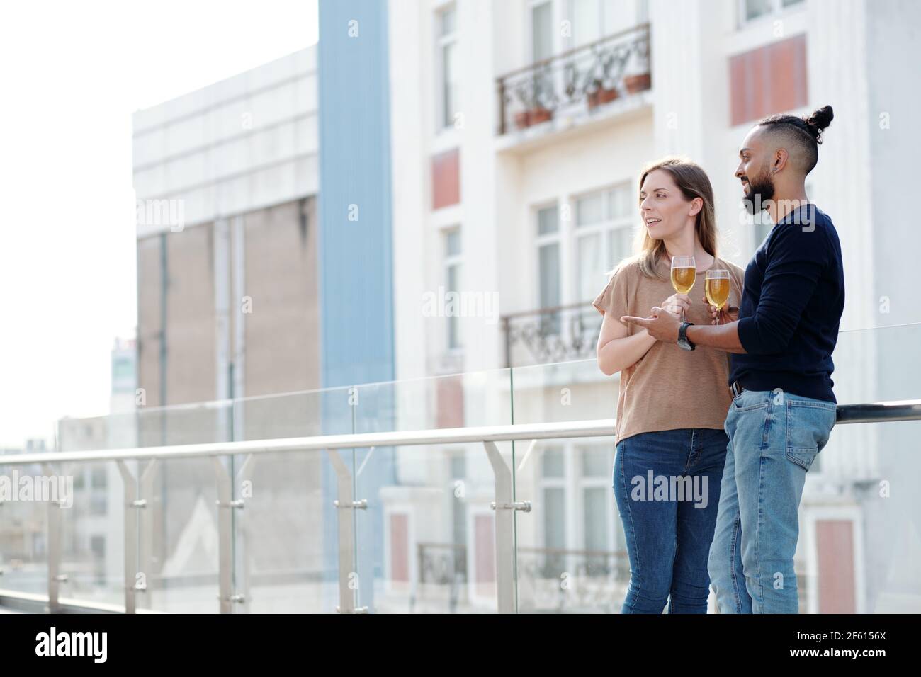 Fröhlicher Freund und Freundin stehen auf dem Dach des Wohnhauses, trinken Weißwein und genießen die Aussicht auf die Stadt Stockfoto