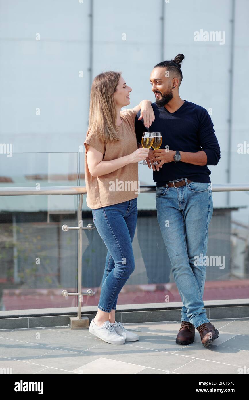 Positives junges Paar, das auf dem Dach steht, Champagner trinkt und sich anschaut Stockfoto