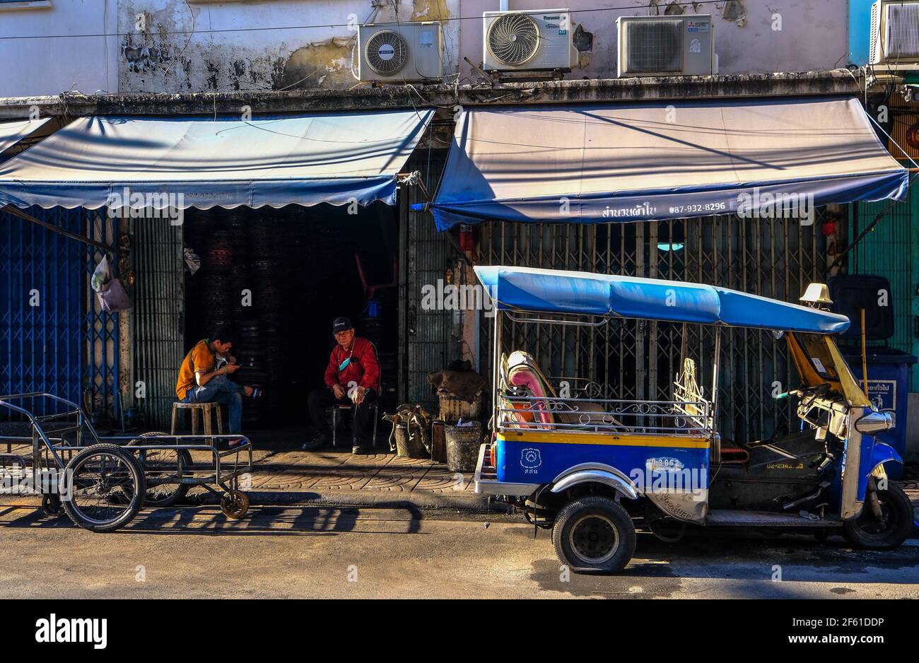 Zwei Männer entspannen sich vor ihrer Werkstatt hinter einem geparkten Tuk-Tuk, das repariert werden muss, in Talat Noi, Bangkok, Thailand Stockfoto