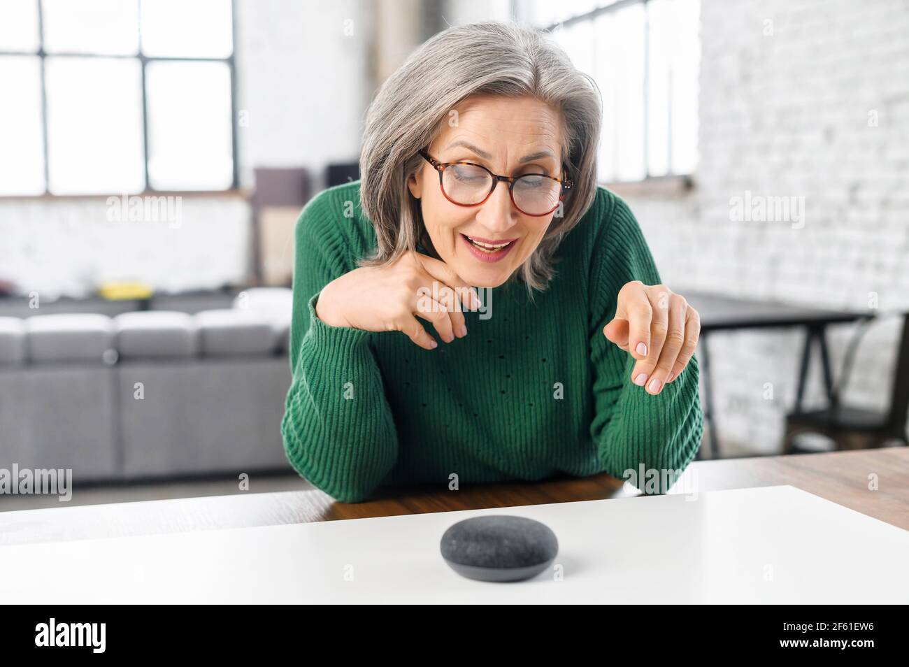 Zeitgenössische pensionierte Frau mit Sprachbefehlen, um intelligente Lautsprecher zu steuern, reife Frau im Gespräch mit dem digitalen virtuellen Assistenten zu Hause, eine Frage stellen oder um Musik zu wechseln Stockfoto