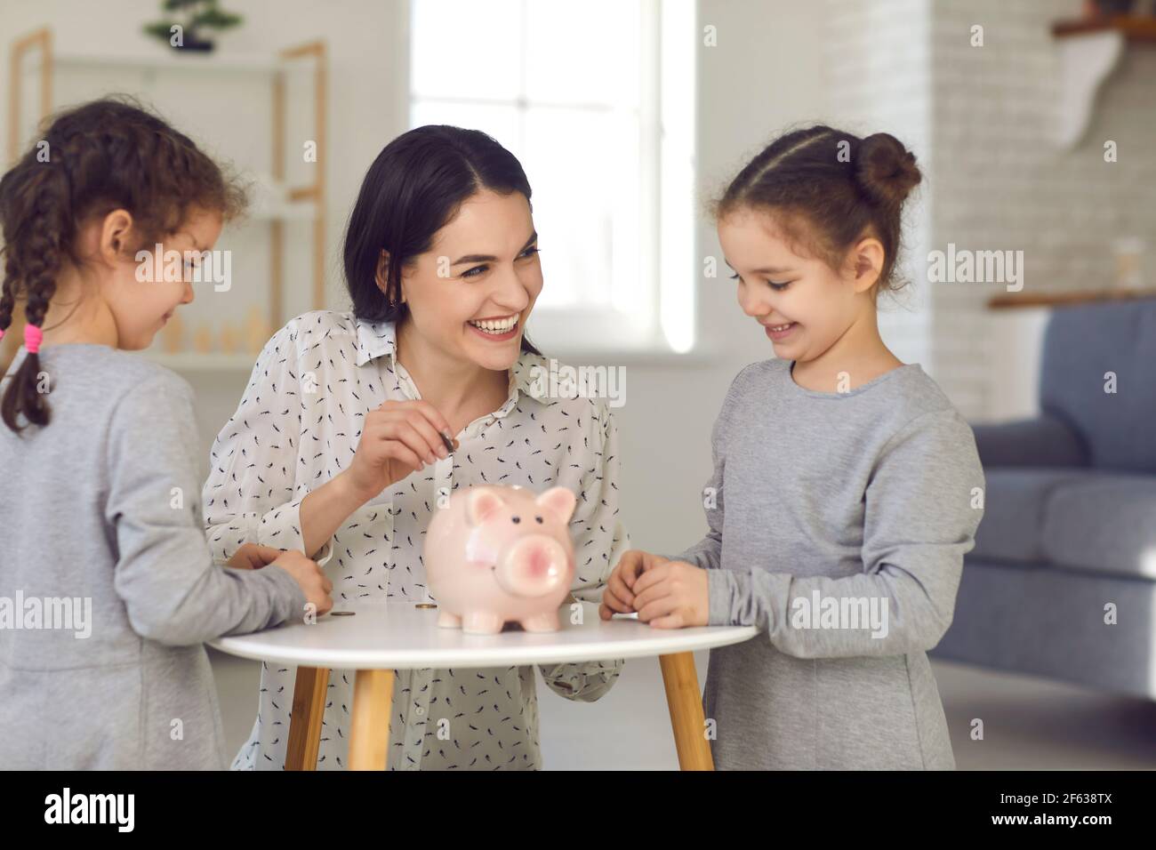 Mama lehrt ihre beiden kleinen Töchter finanzielle Bildung und steckt Geld in eine Sparschwein. Stockfoto