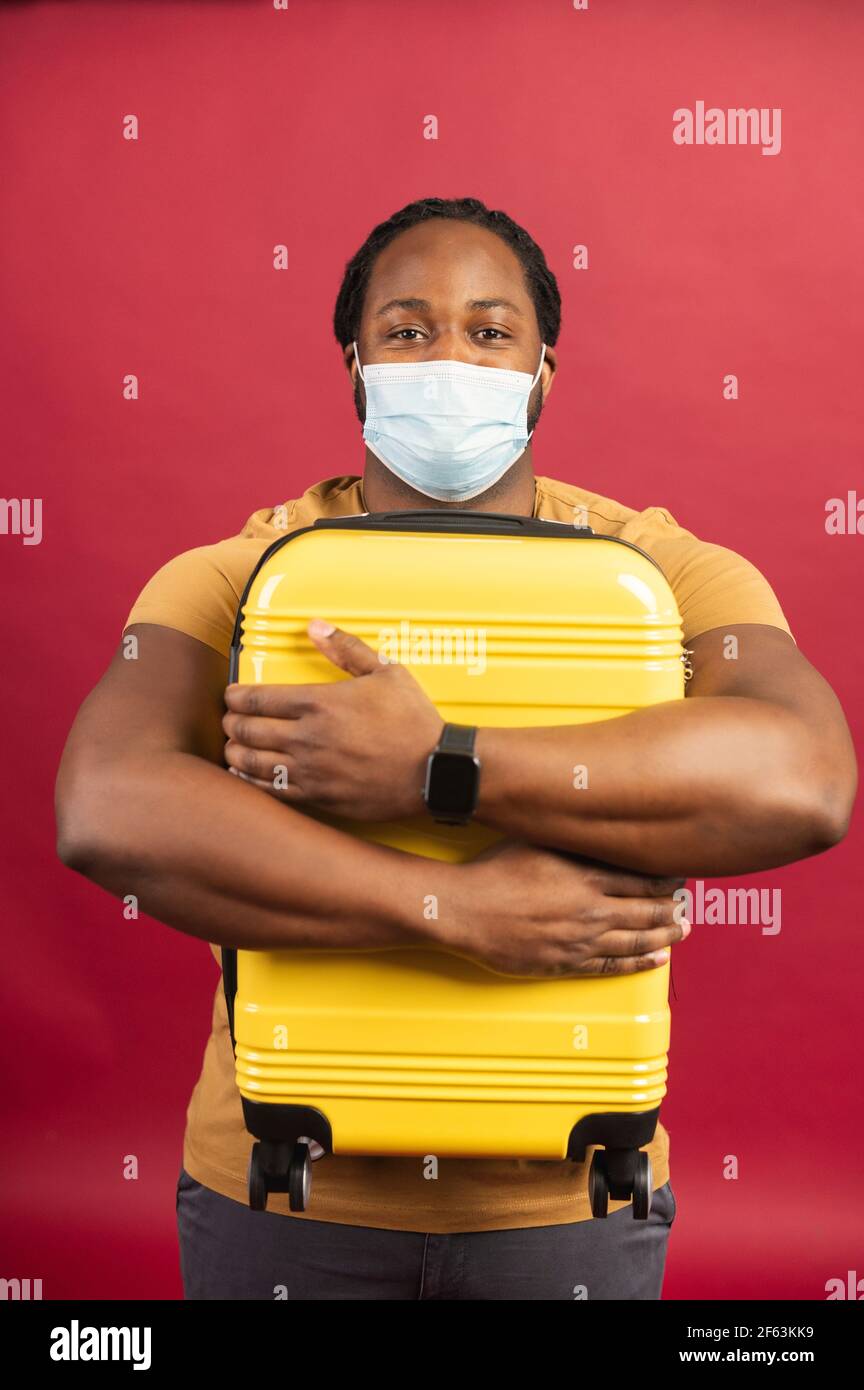 Fröhliches Porträt von afroamerikanischen Kerl trägt schützende medizinische Maske Umarmung einen gelben Koffer. Ein ungeduldiger, um auf eine Reise nach Pandemie Periode schwarzen Mann hält Gepäck gehen, isoliert auf rot Stockfoto