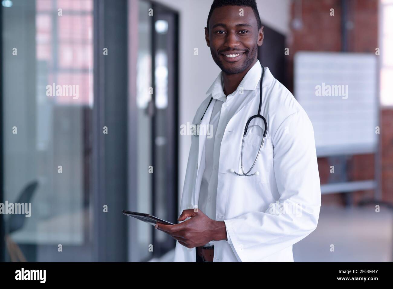 Lächelnder afroamerikanischer Arzt mit weißem Mantel und Stethoskop Mit einem digitalen Tablet Stockfoto