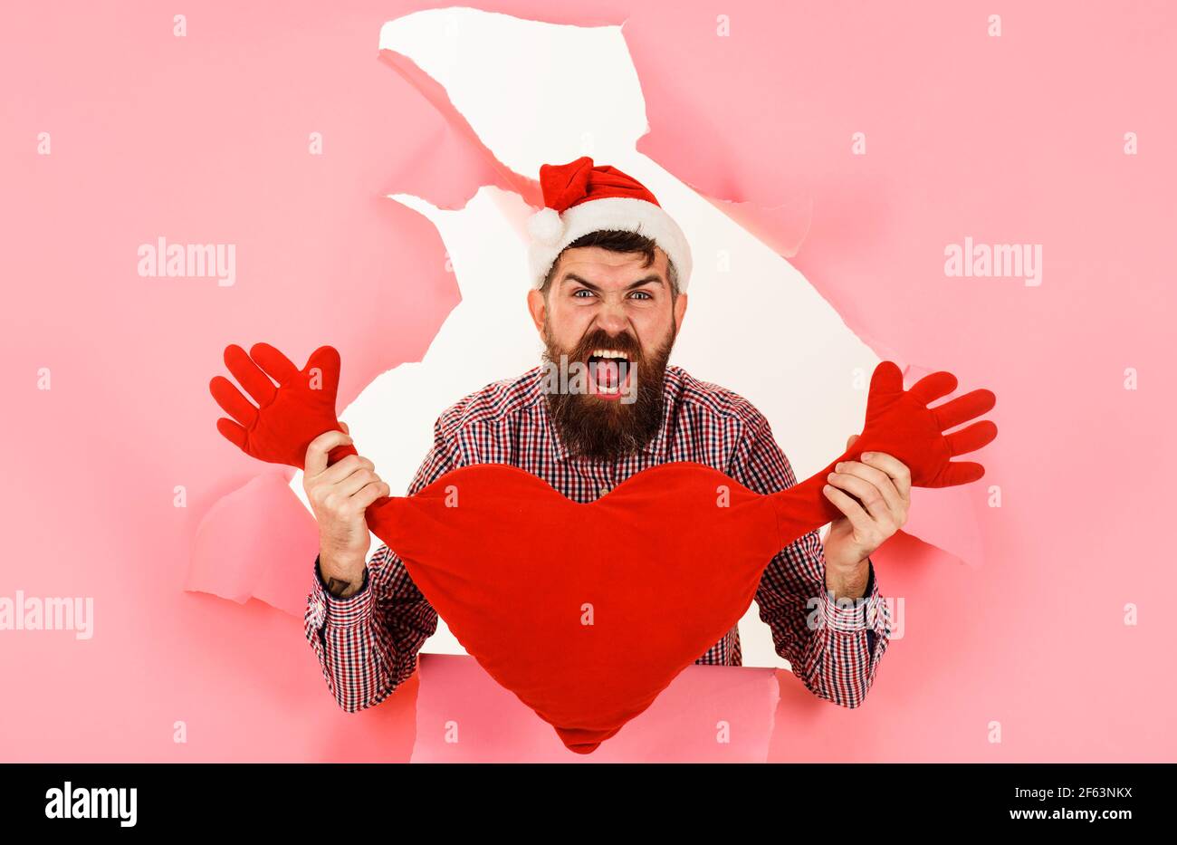 Schreiender Weihnachtsmann mit herzförmigem Kissen. Bärtiger Mann im weihnachtsmütze mit rotem Herzkissen. Stockfoto