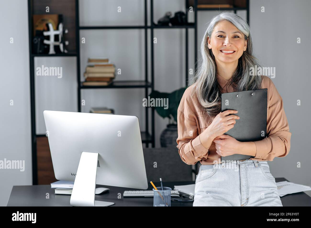 Porträt einer erfolgreichen, grauhaarigen asiatischen Geschäftsfrau mittleren Alters, die im modernen Büro steht, in stilvoller Kleidung gekleidet, Ordner in den Händen hält, die Kamera anschaut und freundlich lächelt Stockfoto