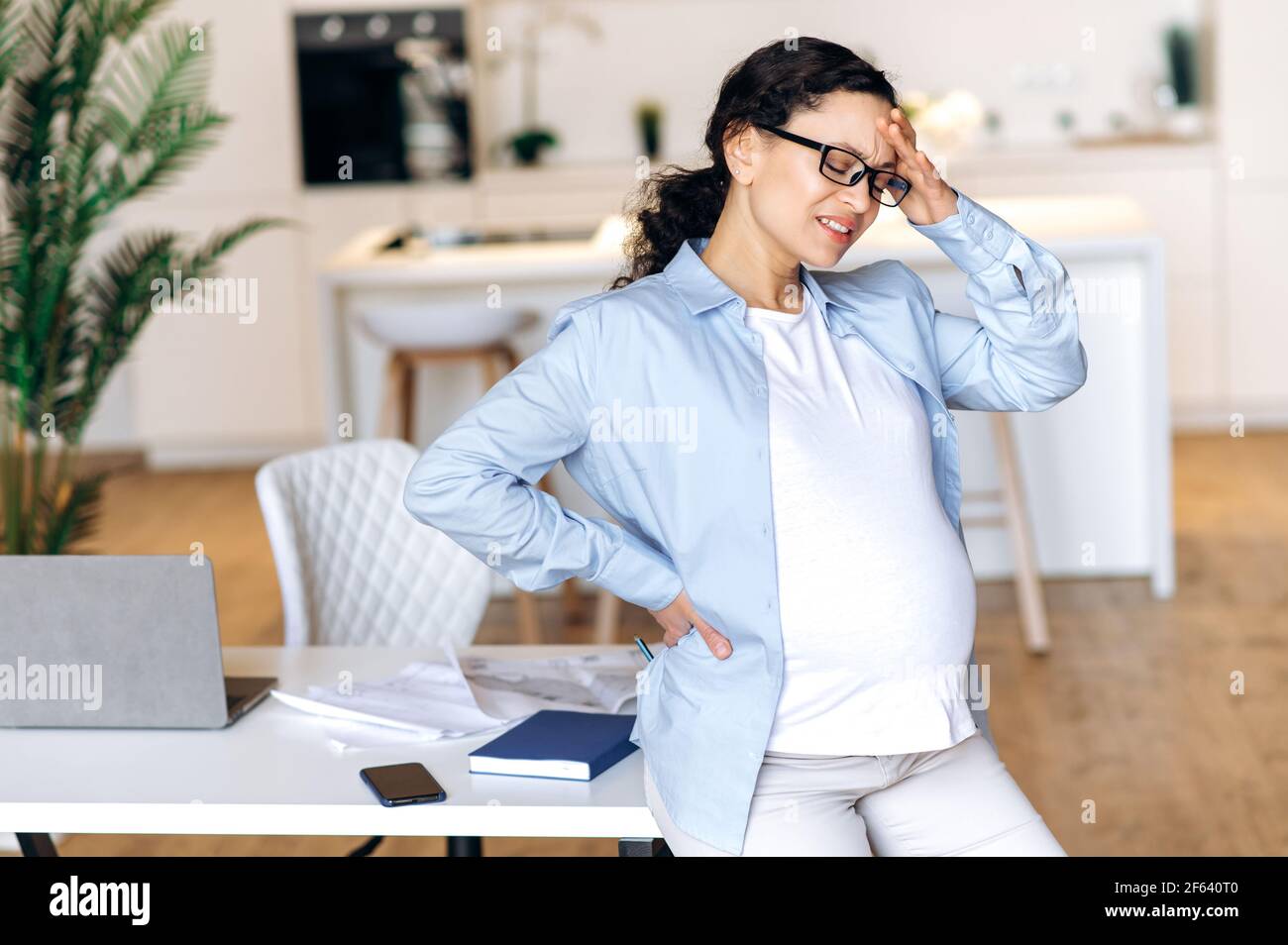 Aufgeregt abgemagert stressig schwanger gemischt Rennen Frau trägt Brille, Geschäftsfrau oder Büroangestellte, steht in der Nähe des Arbeitsplatzes, Pause wegen Kopfschmerzen und Müdigkeit, ihren Kopf mit der Hand halten Stockfoto