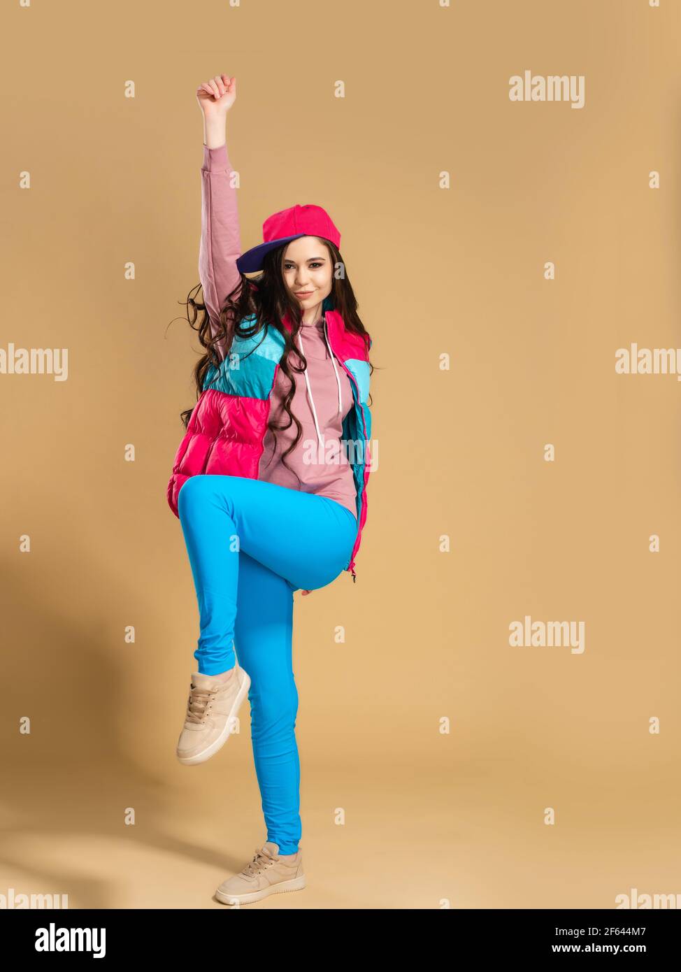 Junges Mädchen in einem rosa Baseball-Cap mit langen Haaren In einer hellen mehrfarbigen Weste und blauen Leggings hebt sie Hand up gegen einen orangen Studio-Backgrou Stockfoto