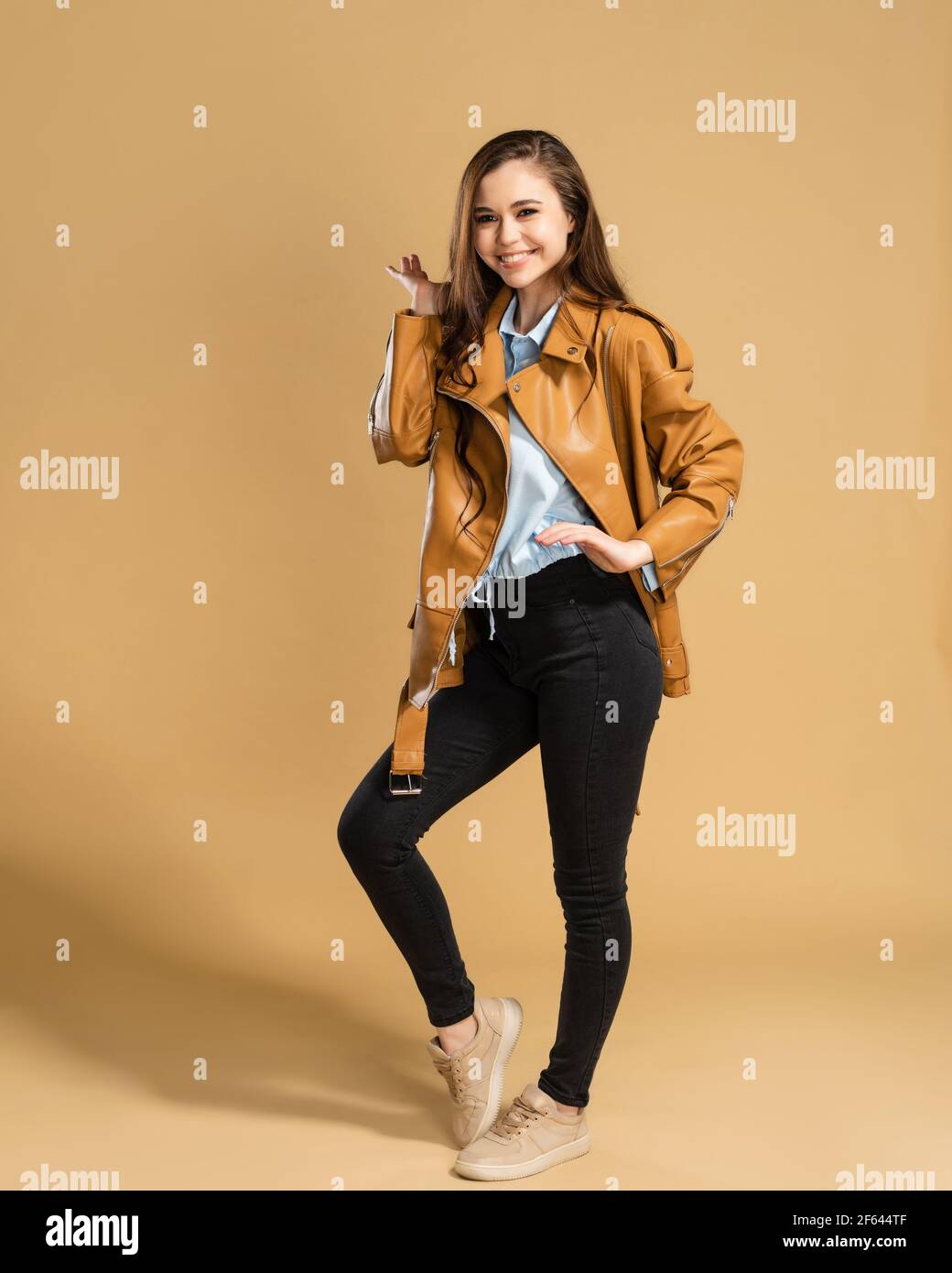 Junge schöne Mädchen mit langen lockigen Haaren in einer braunen Lederjacke posiert auf einem pastellorangen Studio-Hintergrund. Stockfoto