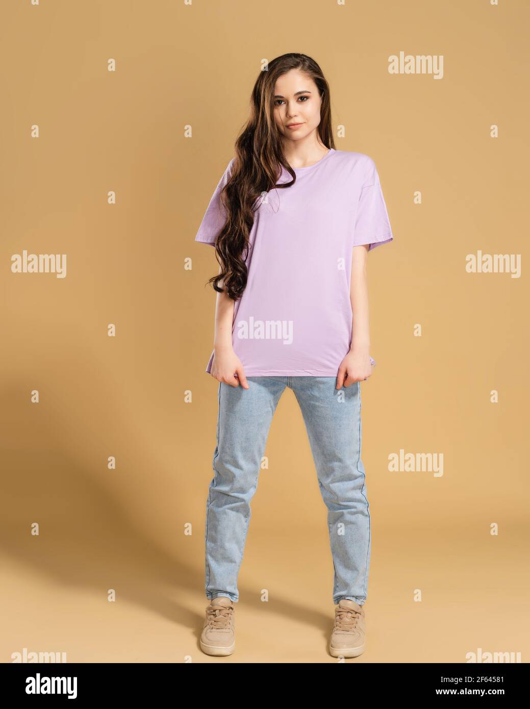 Junge schöne Mädchen mit langen lockigen Haaren in einem lila T-Shirt und blaue Jeans auf einem pastellorange Studio Hintergrund. T-Shirt-Modell. Stockfoto