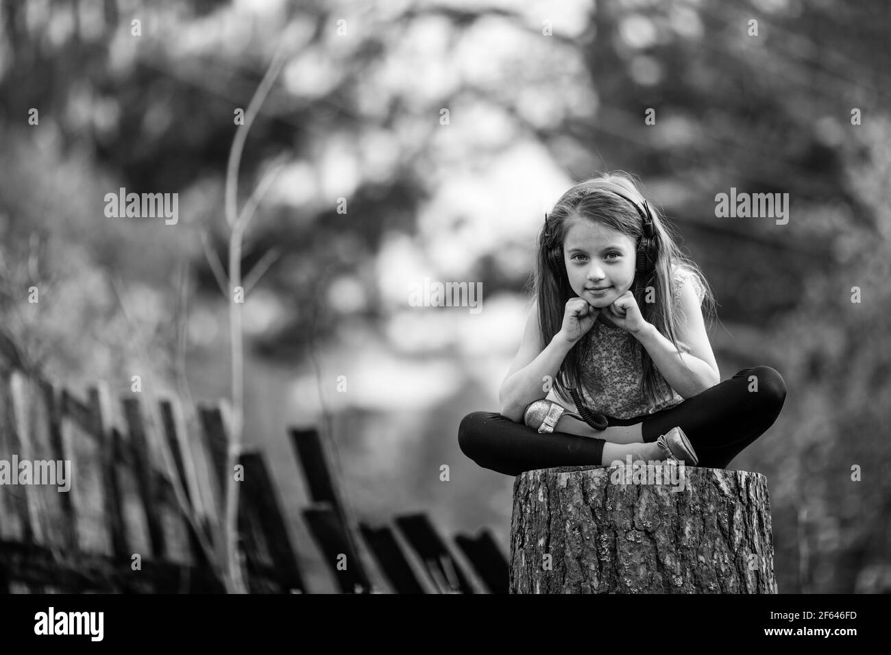 Kleine niedliche Mädchen in Kopfhörer sitzen auf einem Stumpf im Dorf. Schwarzweiß-Foto. Stockfoto