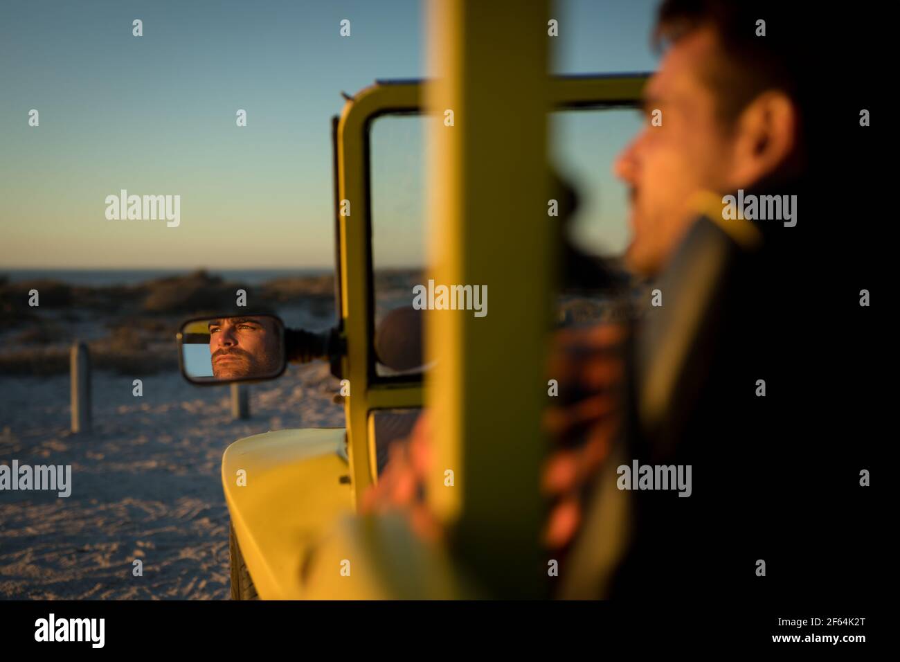 Kaukasischer Mann, der während des Strandbuggys am Meer sitzt Der Sonnenuntergang spiegelt sich im Autospiegel wider Stockfoto