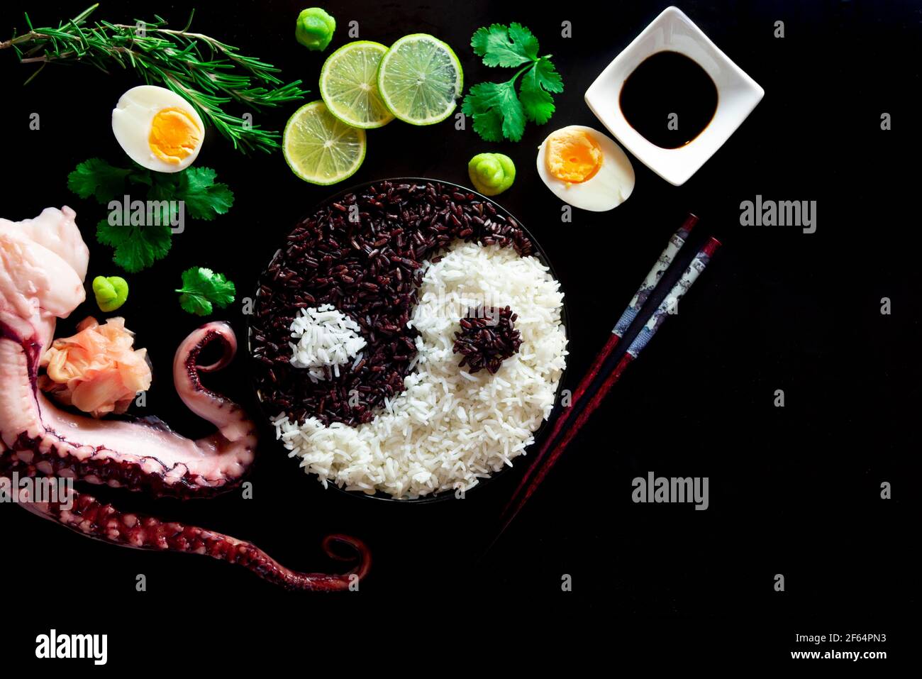Eine Draufsicht auf eine Schüssel mit Yin Yang Reis, Essstäbchen, Limettenscheiben, gekochten Eiern, Sojasauce, Gewürze und Calamari Tentakeln auf schwarzem Hintergrund Stockfoto