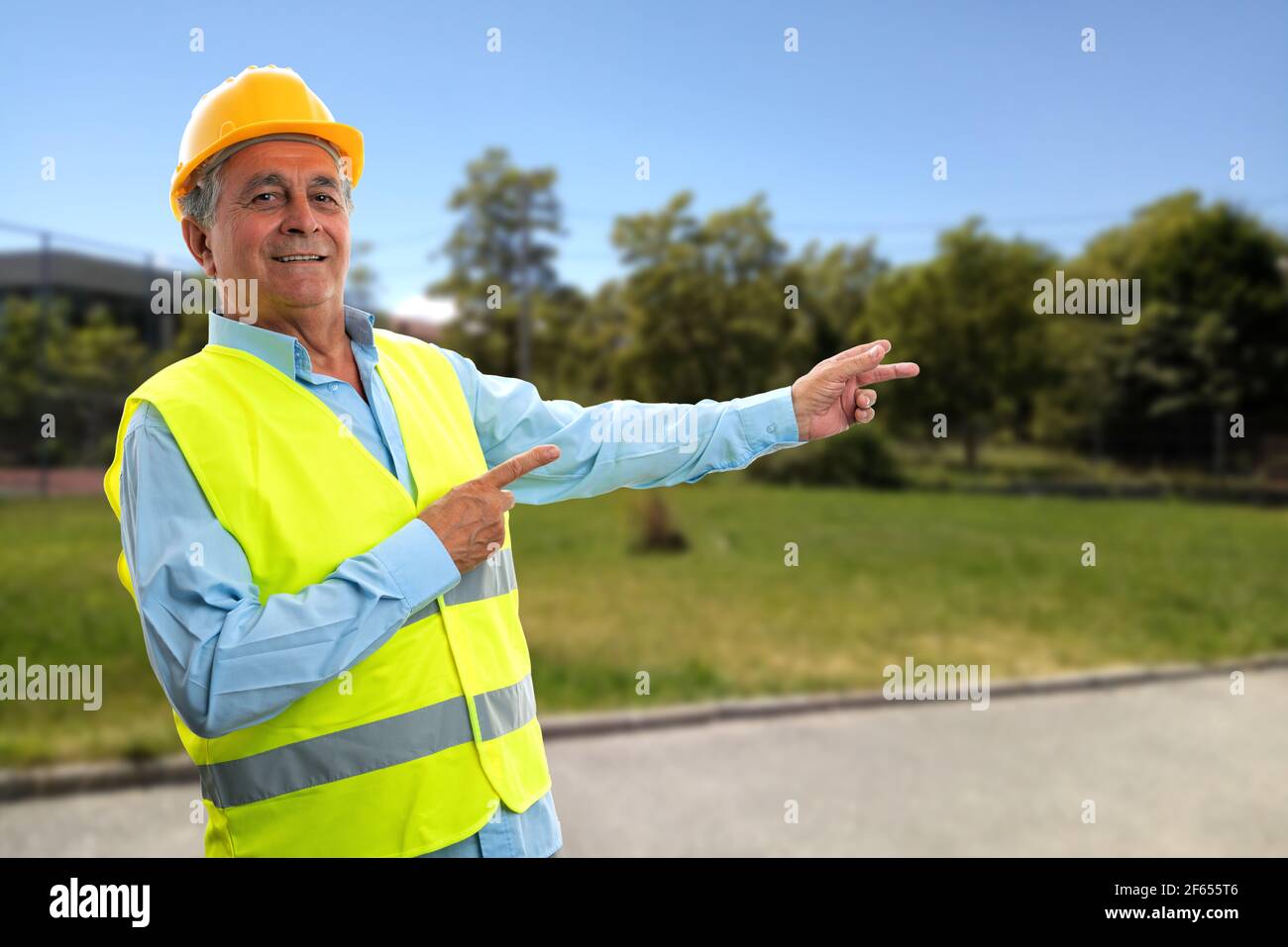 Fröhlicher älterer Konstrukteur Mann lächelnd Zeigefinger auf Park zeigend Natur Hintergrund trägt fluoreszierende Weste und schützende Hardhut Stockfoto