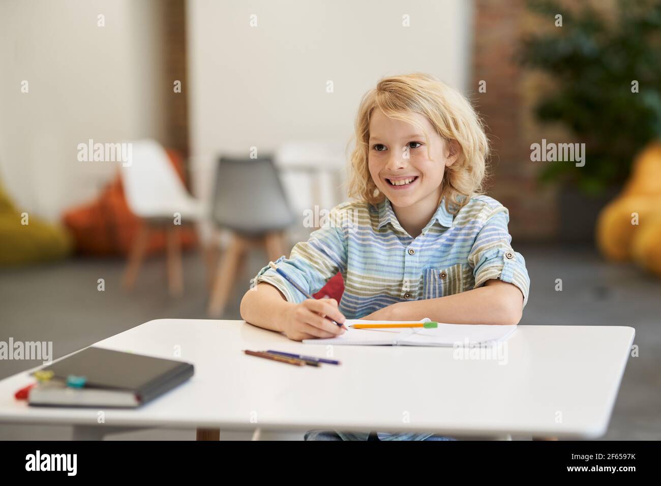 Glücklicher Schüler. Fröhlicher Schuljunge lächelt weg, während er in seinem Notizbuch schreibt und am Schreibtisch im Klassenzimmer sitzt Stockfoto