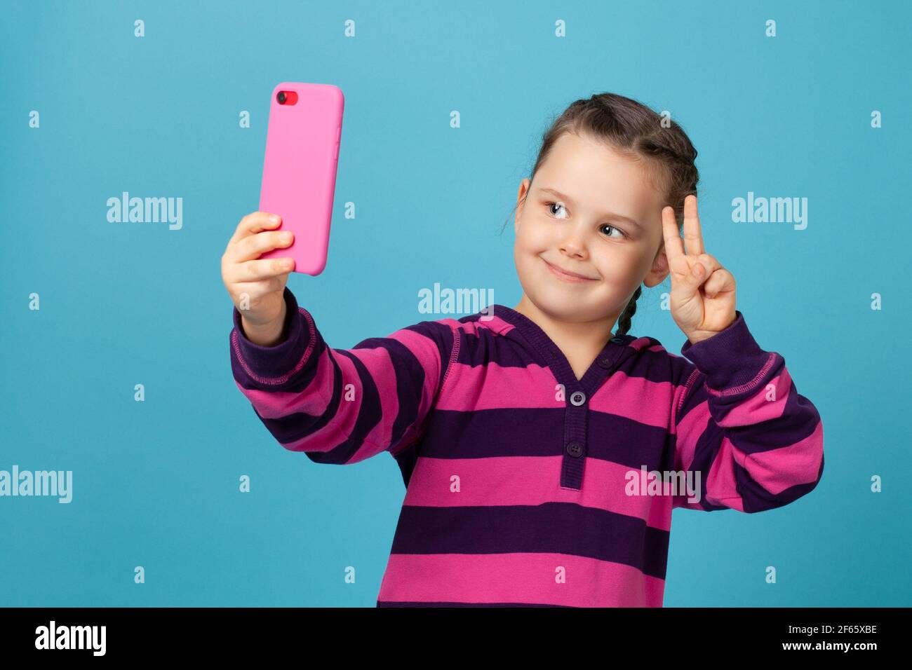 Porträt des Kindes Mädchen lächelnd und Selfie, Telefonanruf auf rosa Telefon und zeigt Siegeszeichen mit Fingern, isoliert auf einem blauen Hintergrund Stockfoto