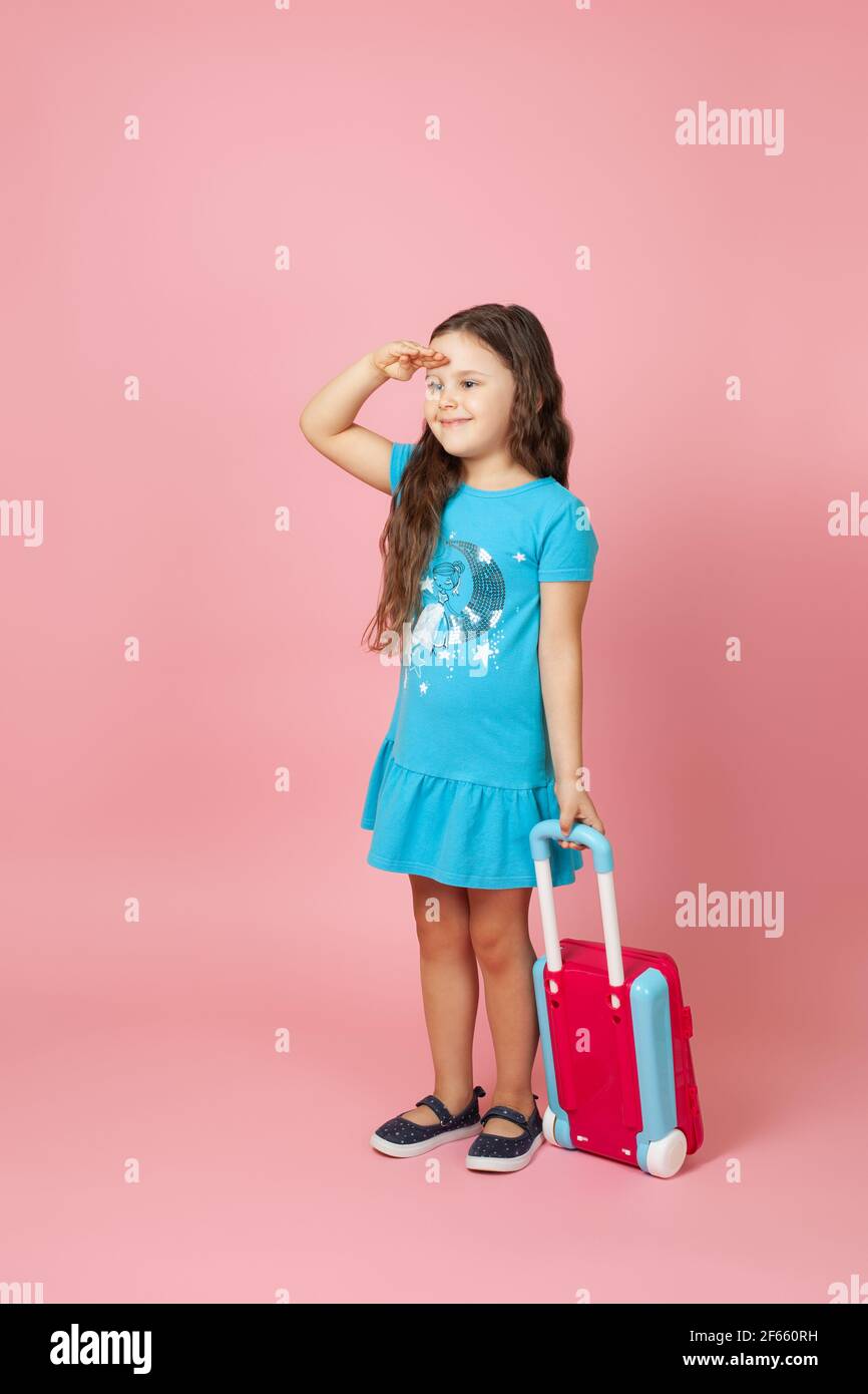 Ganzkörperportrait eines langhaarigen Mädchens, das einen Koffer trägt und mit der Hand über den Kopf weit weg schaut, isoliert auf einem rosa Hintergrund Stockfoto
