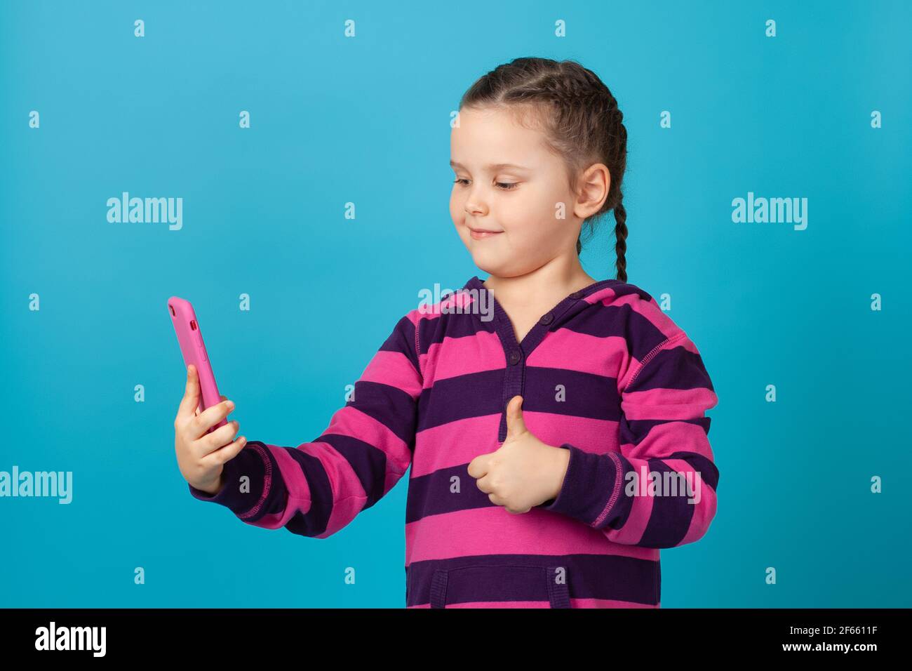 Nahaufnahme eines fünfjährigen Mädchens mit Zöpfen, die auf einer Videotelefonverbindung sprechen und einen Daumen nach oben geben, isoliert auf blauem Hintergrund Stockfoto