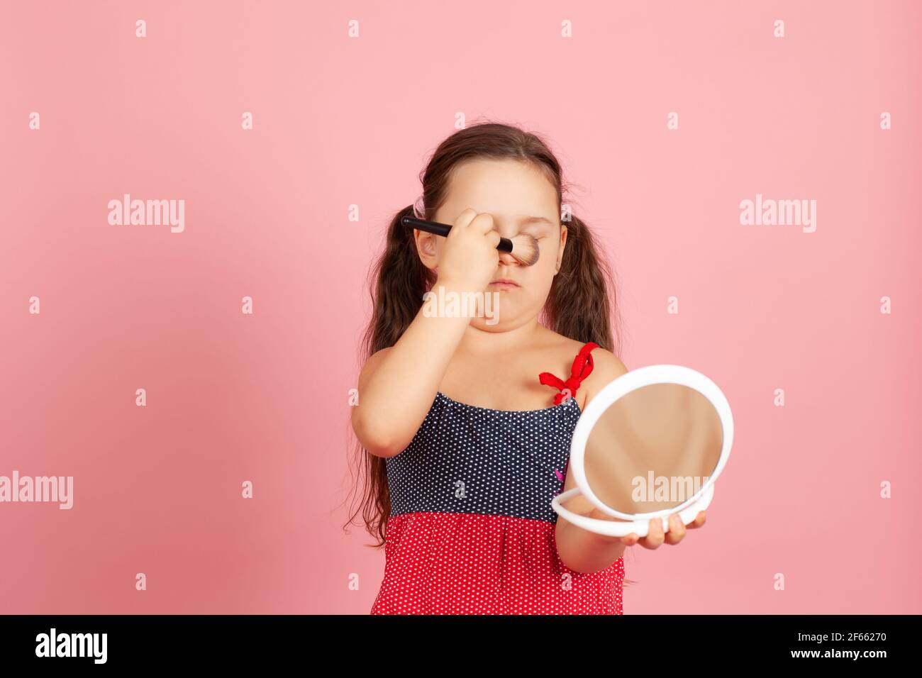 Lustige, ungeschickte fünfjährige Mädchen folgt ihrer Mutter und versucht, mit einem Puderpinsel isoliert auf einem rosa Hintergrund zu schminken Stockfoto