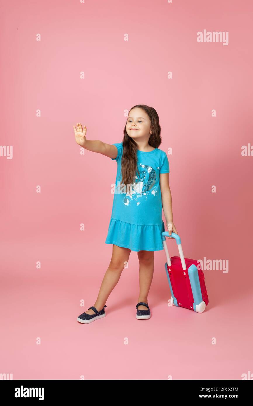 In voller Länge Porträt eines langhaarigen charmanten Mädchen mit einem Koffer und winken eine Grußhand, isoliert auf einem rosa Hintergrund Stockfoto