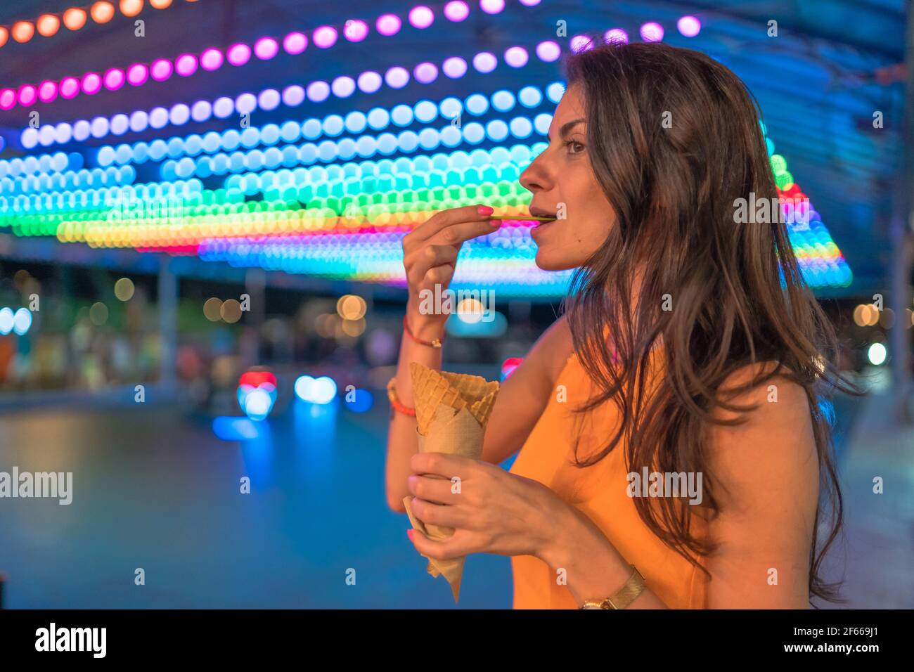 Porträt einer glücklichen Frau, die Eis mit Waffelkegel isst Beim Karneval mit bunten Lichtern im Hintergrund Stockfoto