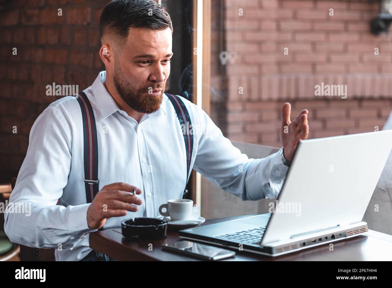 Junger Hipster-Geschäftsmann mit einem Videoanruf in einem Café, während er raucht und Kaffee trinkt Stockfoto