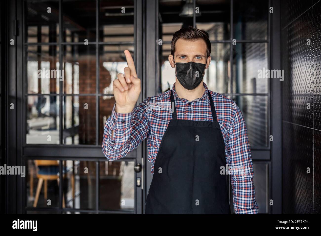 Arbeiten Sie in einem Restaurant während der Krone. Ein Porträt eines männlichen Kellners, der eine Maske mit dem Finger einer Hand trägt, zeigt ein Zeichen einer unbefriedigenden Situation Stockfoto