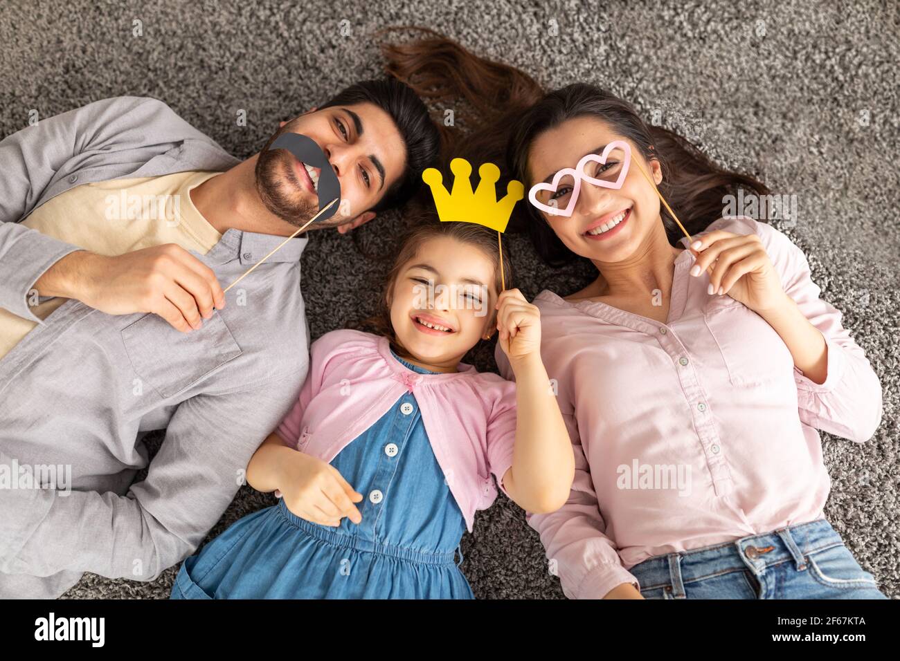 Lustige arabische Vater, Mutter und Tochter Spaß mit Krone, Brille und Schnurrbart auf Stöcken, auf Teppich liegend, Draufsicht Stockfoto