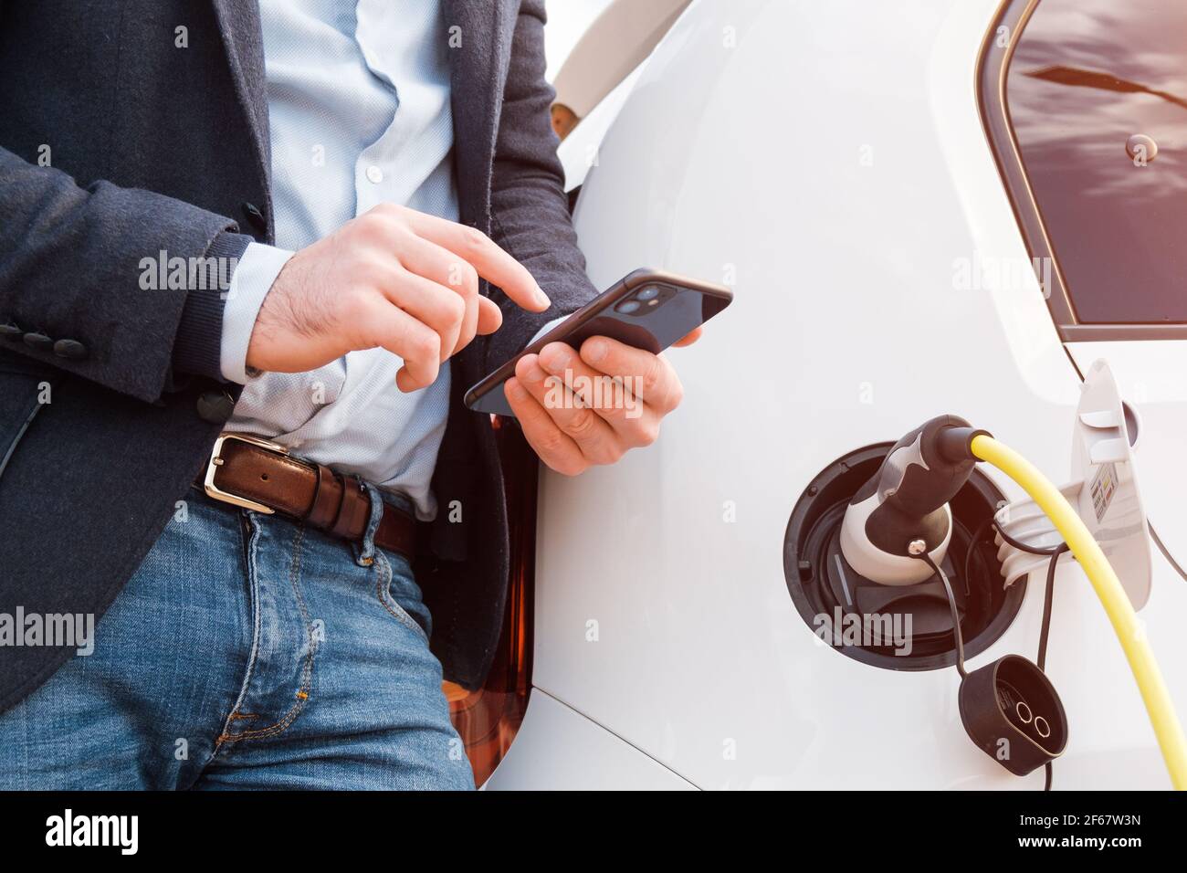 Mann, der in der Nähe des Elektrofahrzeugs oder Elektroautos steht Und mit dem Smartphone bei Sonneneinstrahlung bezahlen Stockfoto