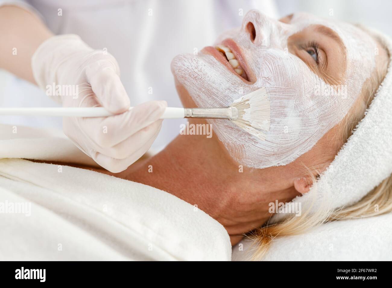 Nahaufnahme Foto der Kosmetikerin Anwendung Ton Gesichtsmaske auf Gesicht einer älteren Frau Stockfoto