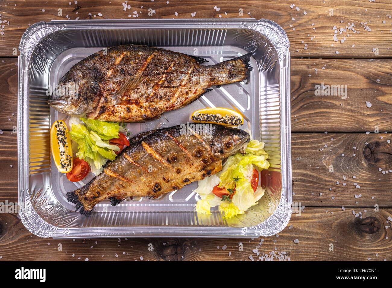 Gegrillter Fisch mit Gemüse, Paprika, grobes Salz, in einem Aluminiumbehälter auf braunem Holzgrund gelegt. Lieferung von Speisen im Restaurant. Stockfoto