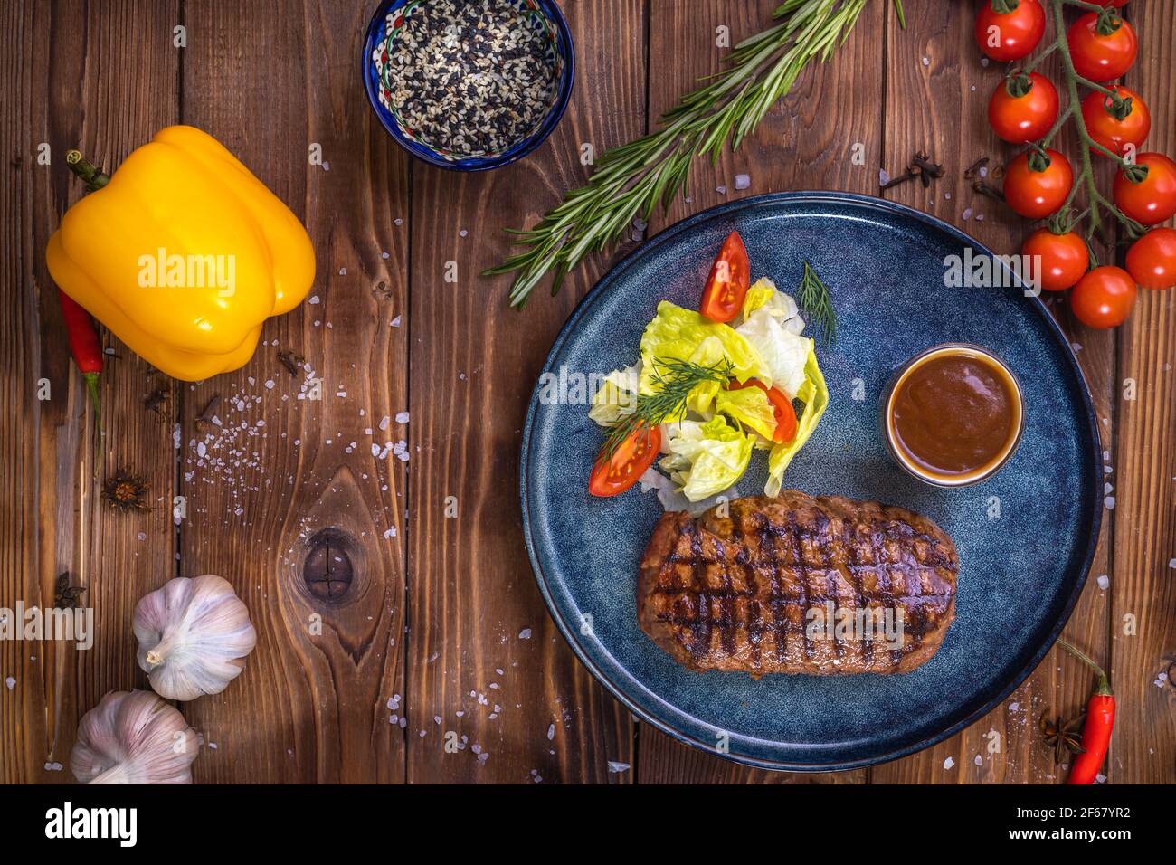 Rinderpastete auf einer Platte mit Gemüse, Salat, Paprika, Rosmarin, Knoblauch und Sesam auf braunem Holzgrund. Restaurant-Service. Stockfoto