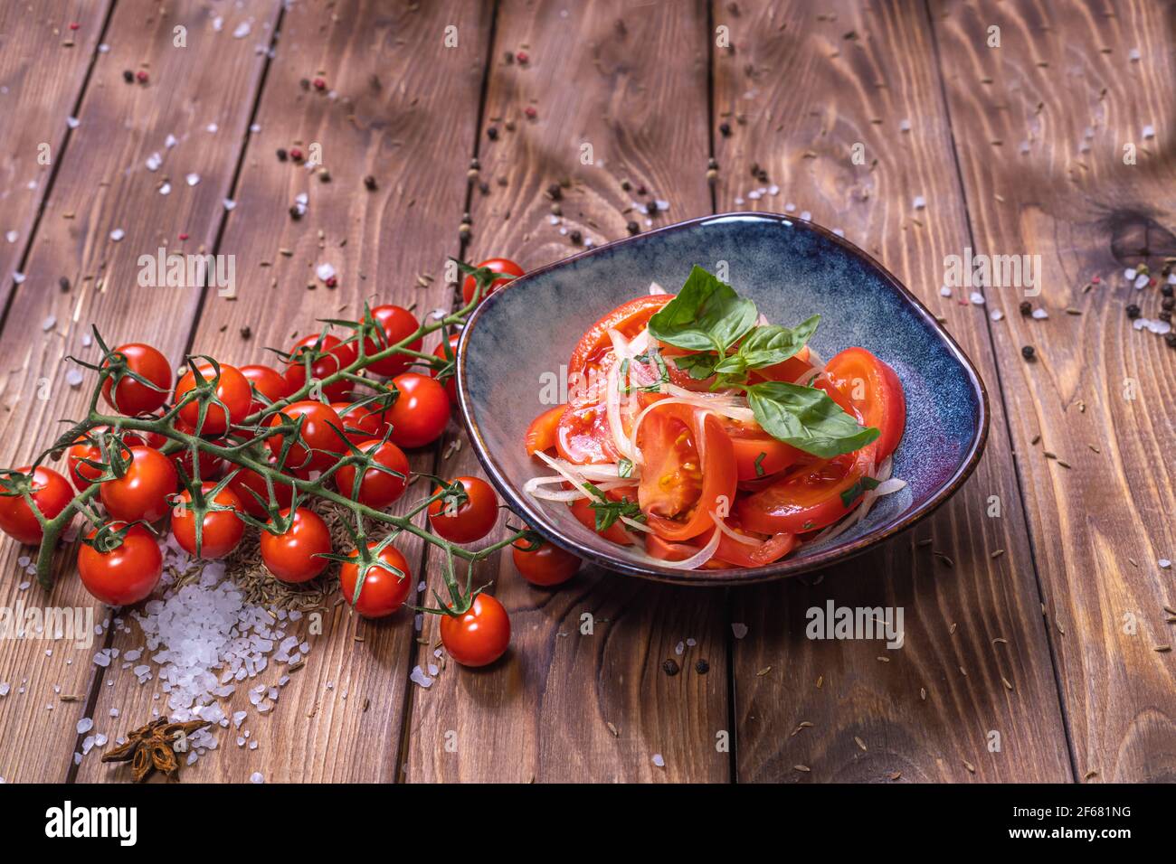 Tomatenscheiben mit Zwiebeln auf einem Teller auf braunem Holzgrund, verziert mit Kirschtomaten, grobem Salz und Pfeffererbsen. Das Konzept von healt Stockfoto