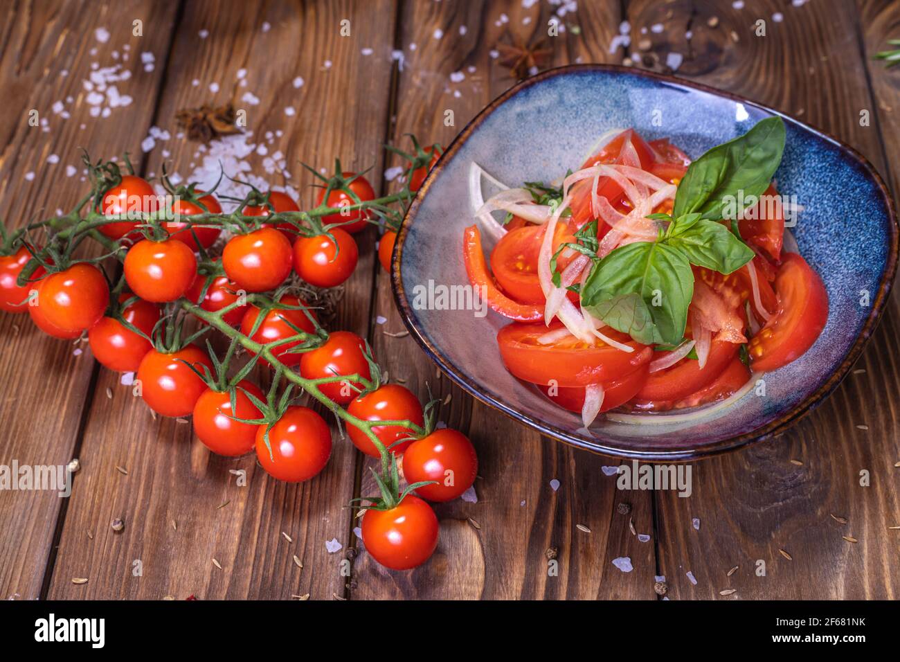 Tomatenscheiben mit Zwiebeln auf einem Teller auf braunem Holzgrund, verziert mit Kirschtomaten, grobem Salz und Pfeffererbsen. Das Konzept von healt Stockfoto