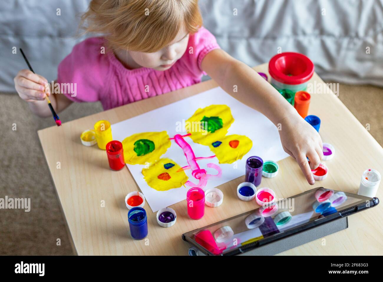 Kleines Mädchen malen auf Papier mit bunten Farben, Blick auf Tablette, sitzen am Tisch zu Hause. Online-Schulkonzept. Stockfoto