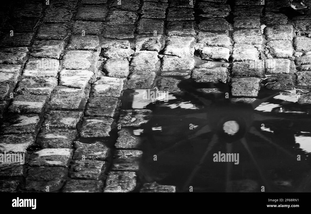 Das hölzerne Rad eines Pferdewagens spiegelt sich nach dem Regen in einer Wasserpfütze wider. Schwarzweiß-Foto. Stockfoto