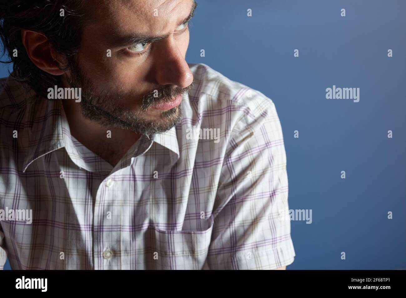 Junger, hübscher, ernsthafter bärtiger Mann mit gestreiftem Hemd, der vor hellblauem Hintergrund sitzt und wegschaut. Denken an die Zukunft, Konzept der pl Stockfoto