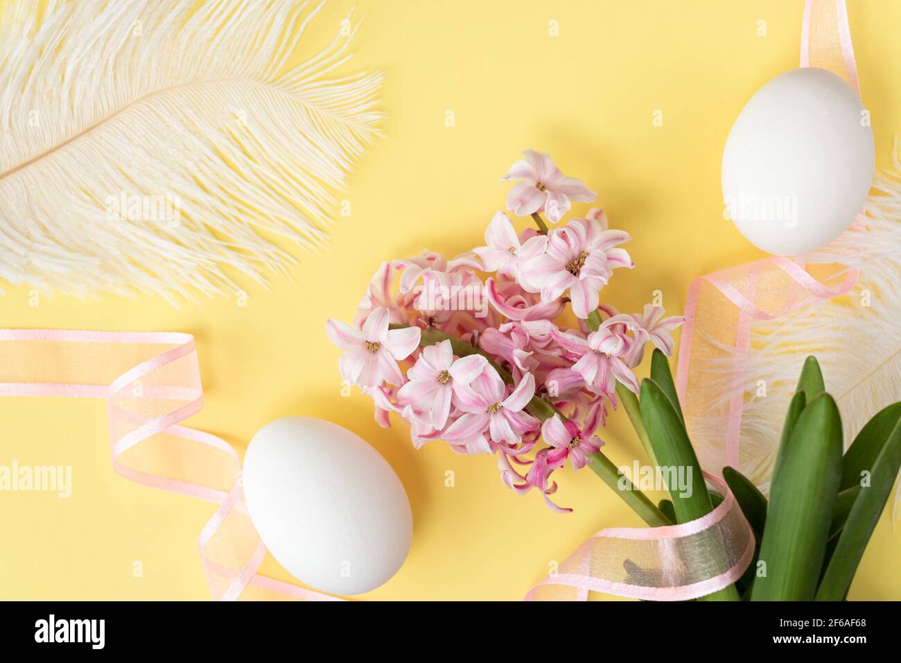 Hyazinthen Blüten mit zwei weißen Eiern, weißen Federn und rosa Band auf pastellgelben Farben. Frohe Ostern Konzept. Osterhintergrund. Draufsicht Stockfoto