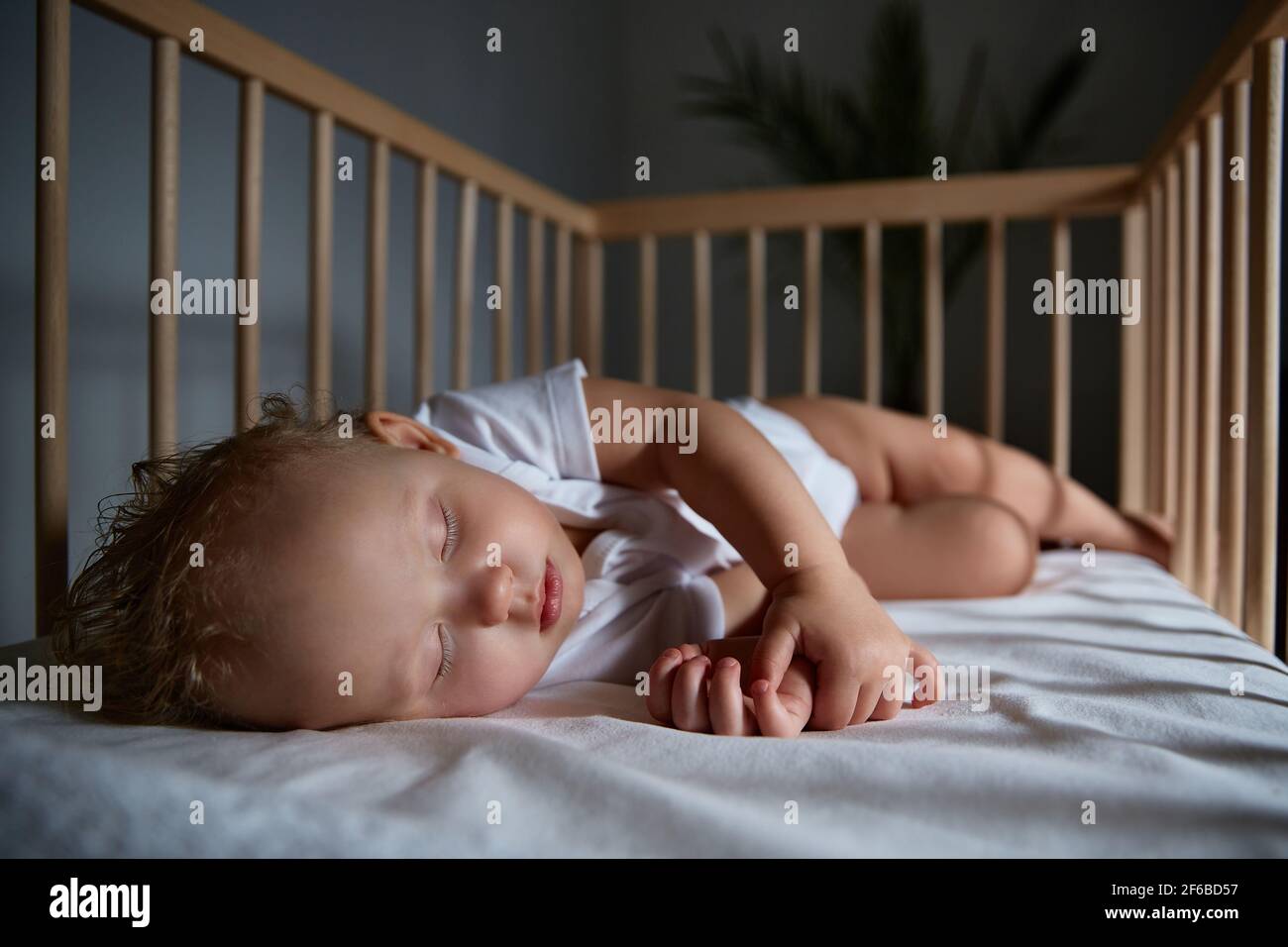 Nettes Baby schläft nachts in einer Wiege für Babys ohne Baldachin und Stoßstangen. Sicher schlafen in einem dunklen, geräumigen Zimmer mit einem Kinderbett und einer Blume. Innen o Stockfoto