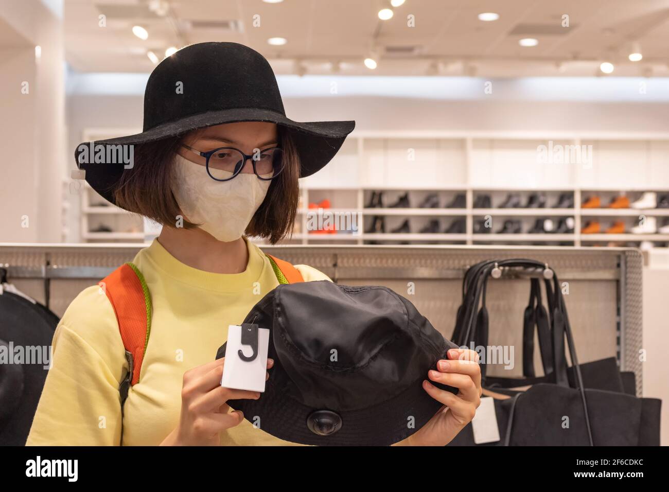 Ein schönes junges Mädchen in einer Schutzmaske wählt während des Rabattzeitraums einen Hut im Laden. Verschiedene Accessoires für einen stilvollen Look. Safe Stockfoto