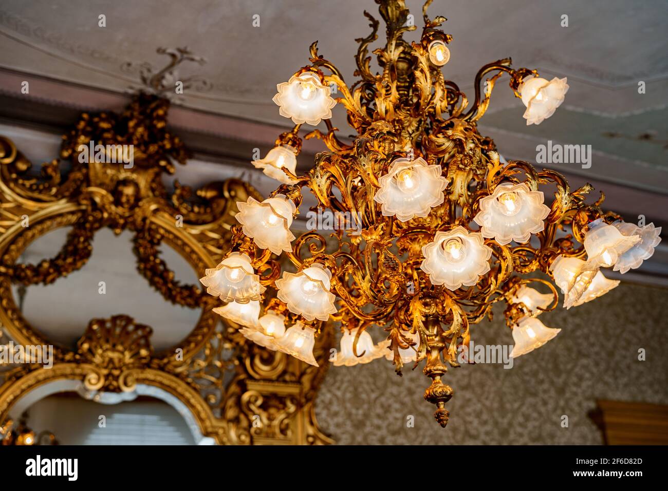 Großer goldener Kronleuchter mit floralen Farbtönen und floristischen Glühbirnen. Vor dem Hintergrund eines antiken Spiegels mit einem geschnitzten vergoldeten Rahmen. Stockfoto