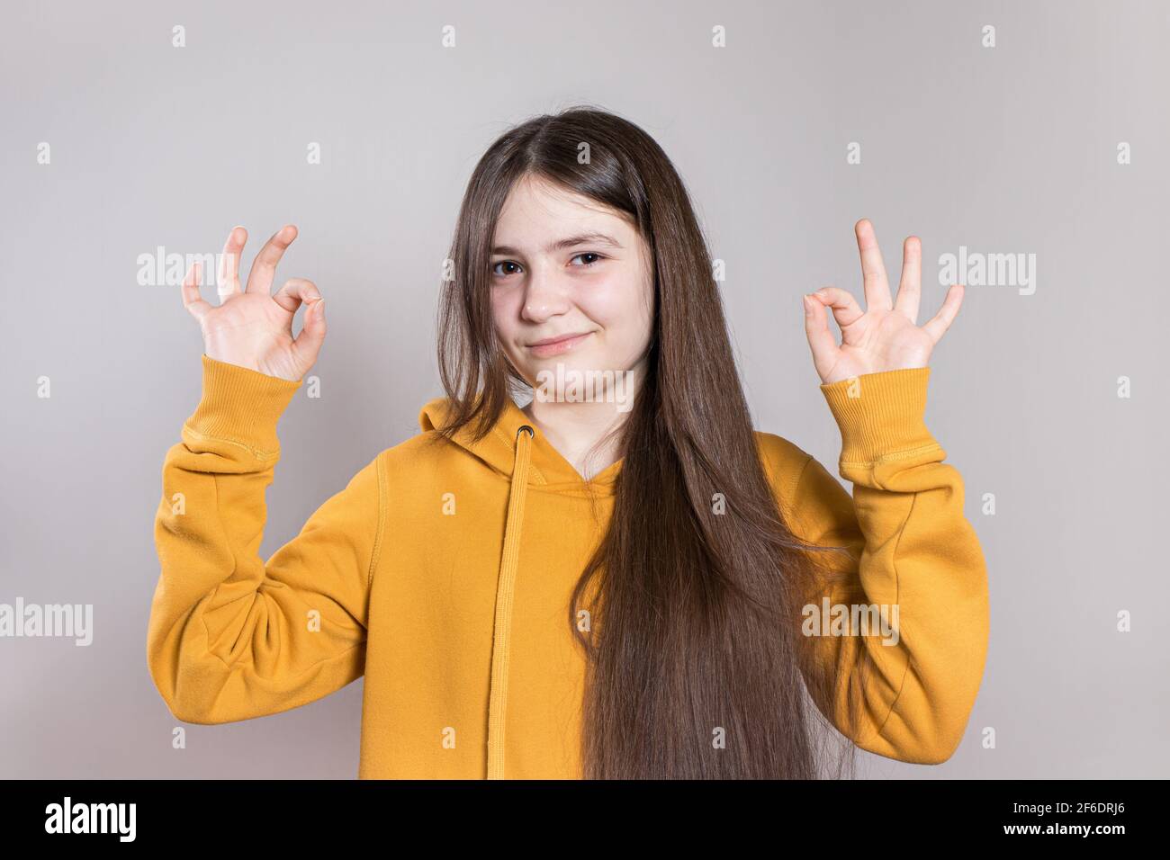 Ein schönes Teenager-Mädchen mit langen Haaren zeigt das Finger-Zeichen OK in einem hellen Hoodie. Kopieren Sie den Platz für Text Stockfoto