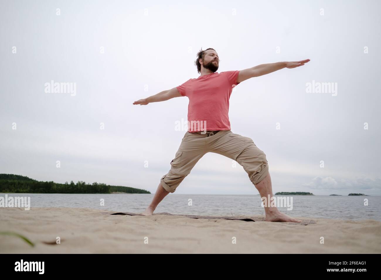 Kaukasischer Mann praktiziert Yoga, er führt virabhadrasana Übung, auf einem Teppich stehen, auf einem Sandstrand am See. Stockfoto