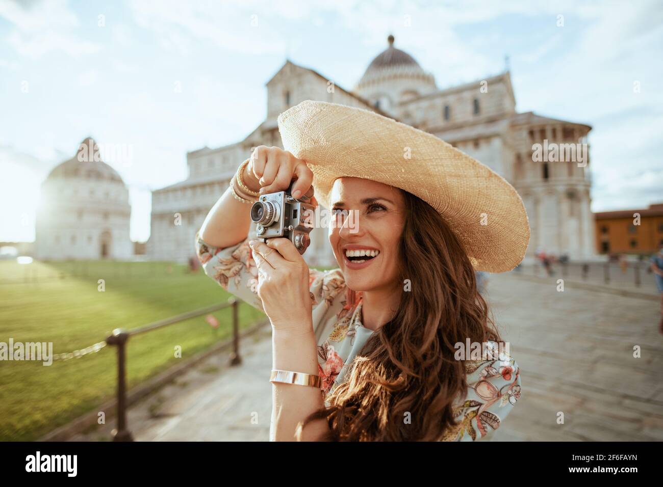 Lächelnde moderne Frau in Blumenkleid mit Retro-Kamera und Hut auf der piazza dei miracoli in Pisa, Italien. Stockfoto