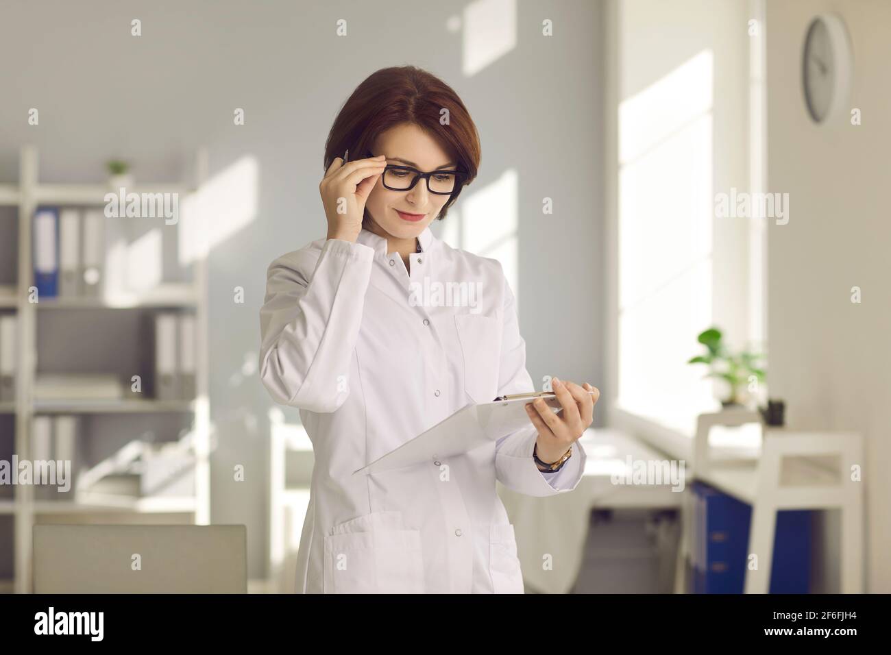 Professionelle Ärztin mit Brille, die die ärztliche Karte des Patienten sorgfältig untersucht. Stockfoto