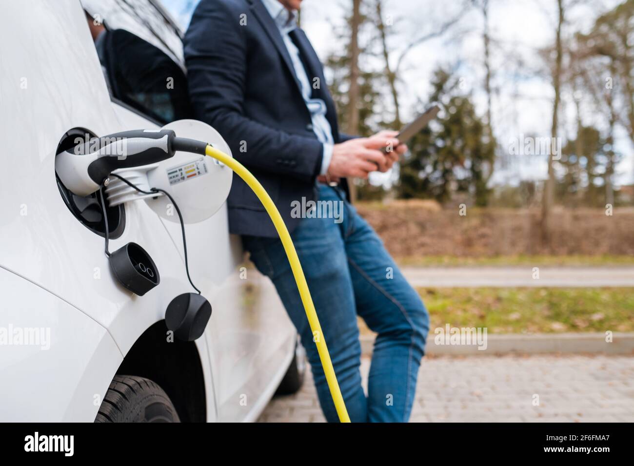 Geschäftsmann, der in der Nähe eines Elektroautos oder Elektroautos steht und auf der Straße ein Tablet benutzt. Stockfoto