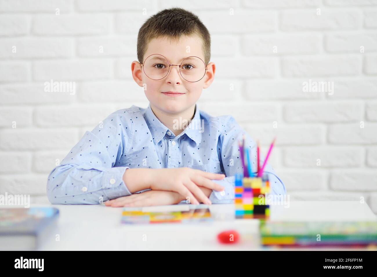 Ein Schuljunge mit Brille, der an einem weißen Tisch sitzt, lächelt vor dem Hintergrund von Notizbüchern und Büchern. Stockfoto