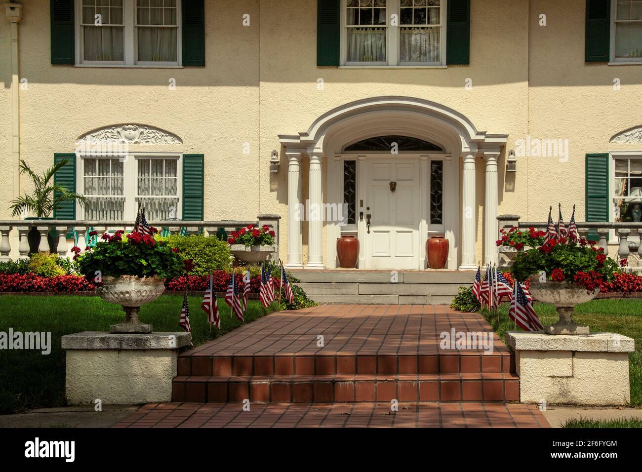 Klassischer gehobener Hauseingang mit hübschen grünen Fensterläden und Säulen Auf der Veranda und dem gemauerten Gang mit Geranien und USA-Flaggen Curb-Look Stockfoto