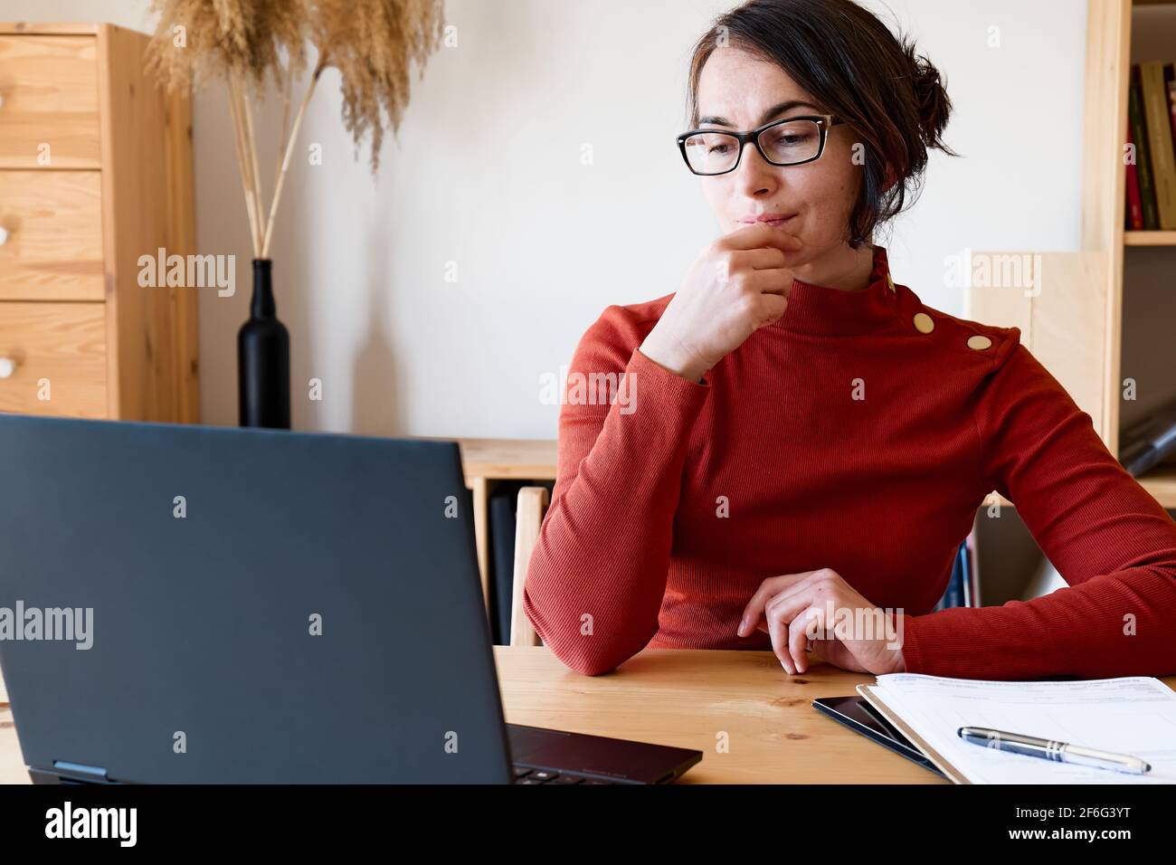 Frau mit rotem Rollkragen und Brille arbeitet mit Laptop in der Nähe des Fensters. Remote Working-Konzept. Stockfoto