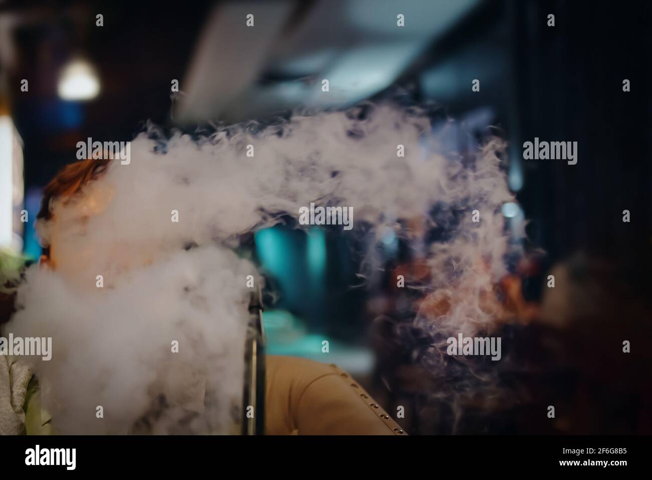 Junger Mann im Café raucht Shisha. Dicker Rauch von Shisha. Mann auf dem Sofa im Restaurant. Neonschilder auf dem Hintergrund. Entspannen Sie sich in der Shisha Bar. Stockfoto