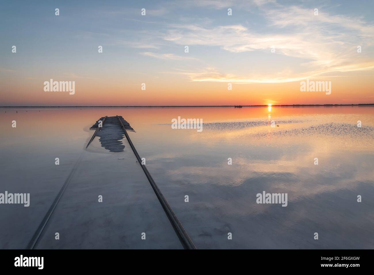Erstaunlicher Sonnenuntergang über einem Salzsee mit wunderschönen Wolken, Sonne, Zug am Horizont, Reflexionen im Wasser und Schienen im Vordergrund. Lake Bursol, EIN Stockfoto